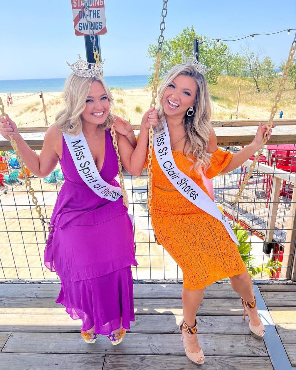 two miss michigan scholarship program contestants sit on swings at the deck restaurant. both are blonde, smiling and wearing sashes stating miss spirit of the state and miss st. claire shores. one wears a purple dress and the other orange. the lake michigan beach sets the backdrop