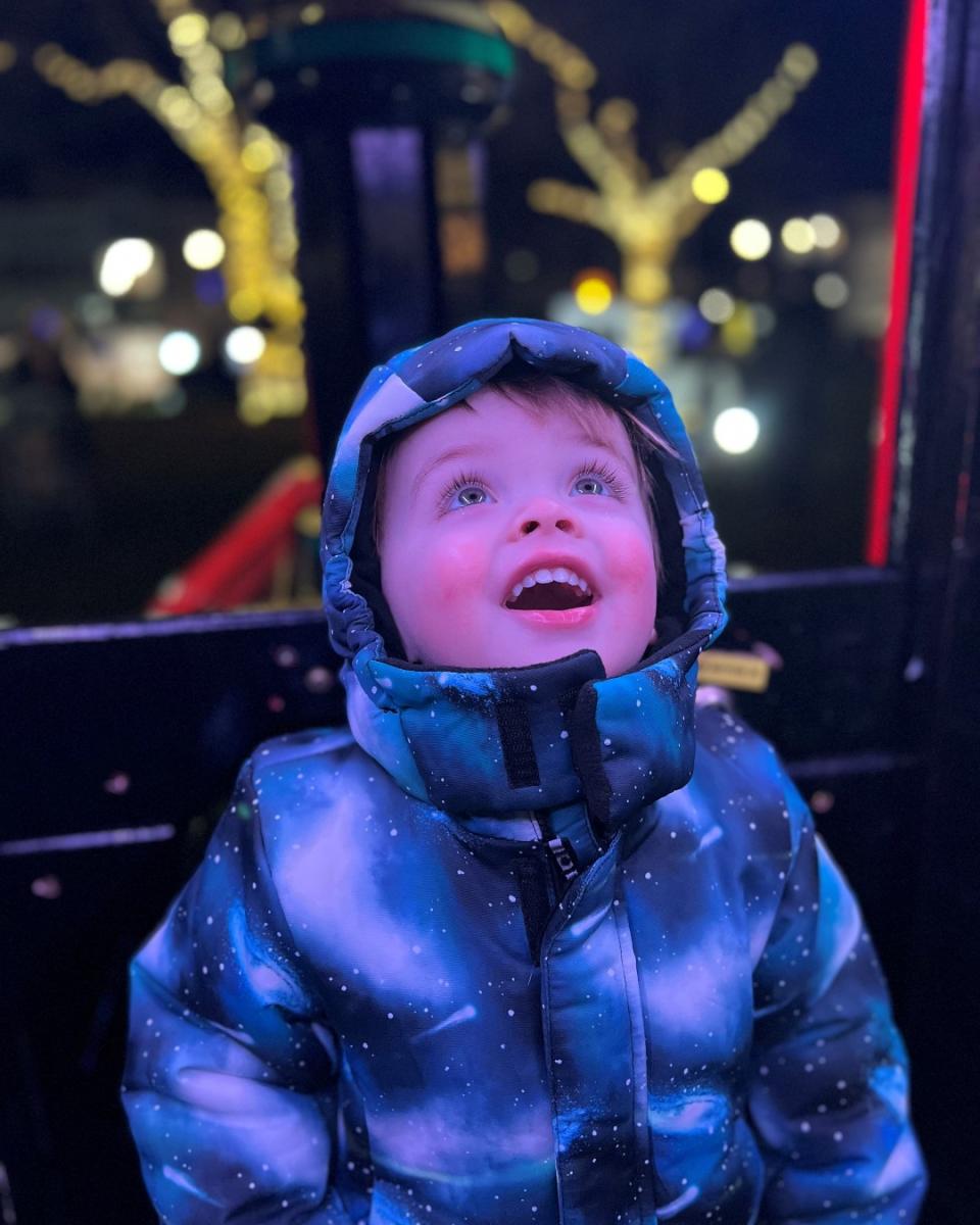 A child wearing a starry winter jacket and a hood looks up in awe at a brightly illuminated scene, possibly at a festive event. The background features blurred lights and decorated trees.