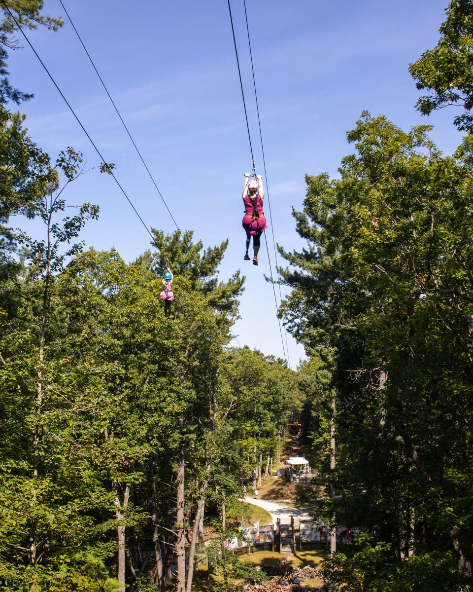 Two individuals in harnesses sail on zipline wires through the dense forest at the Muskegon Luge Adventure Sports Park.
