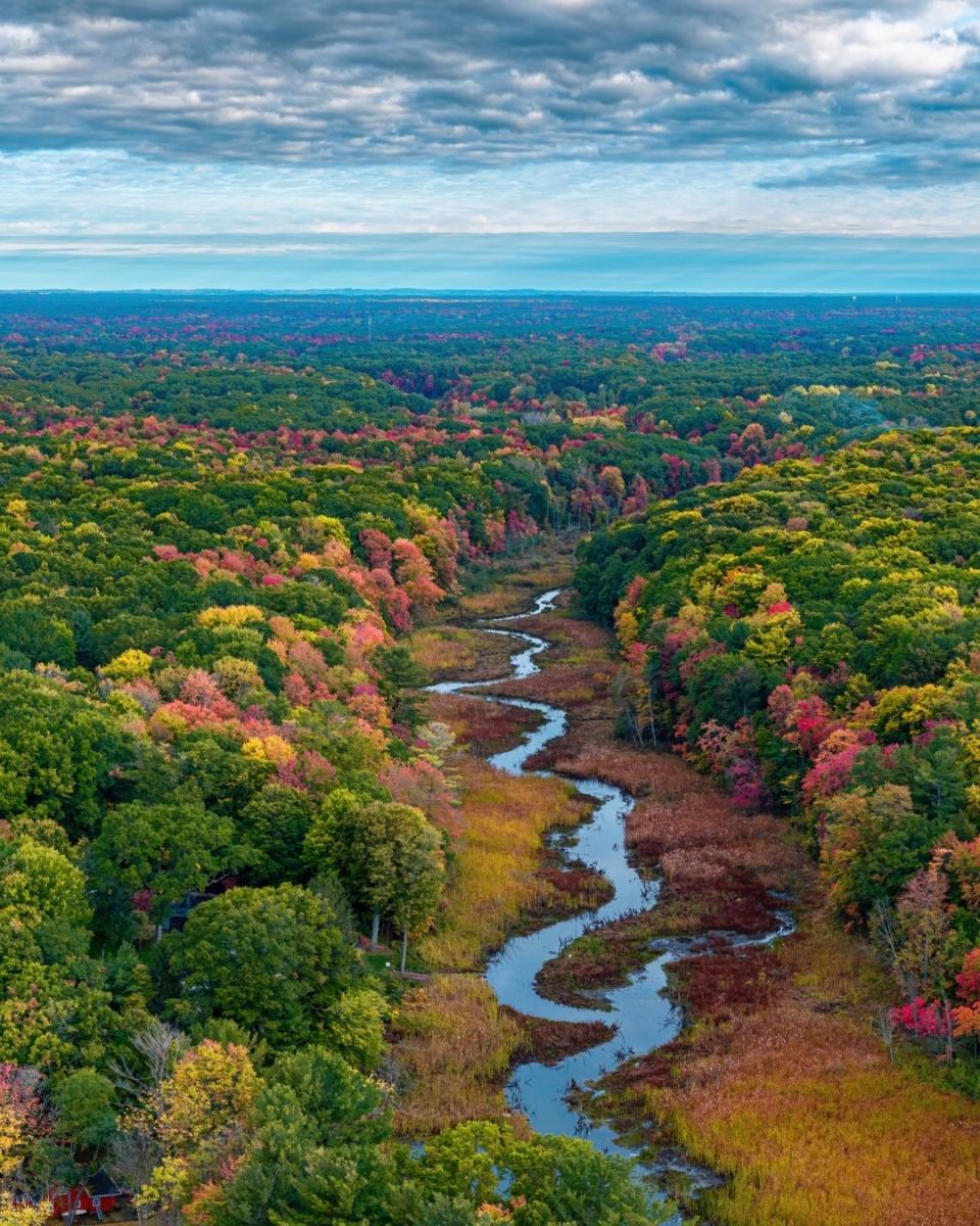 Aerial view of a winding Green Creek in North Muskegon cutting through an autumn forest under a cloudy sky.