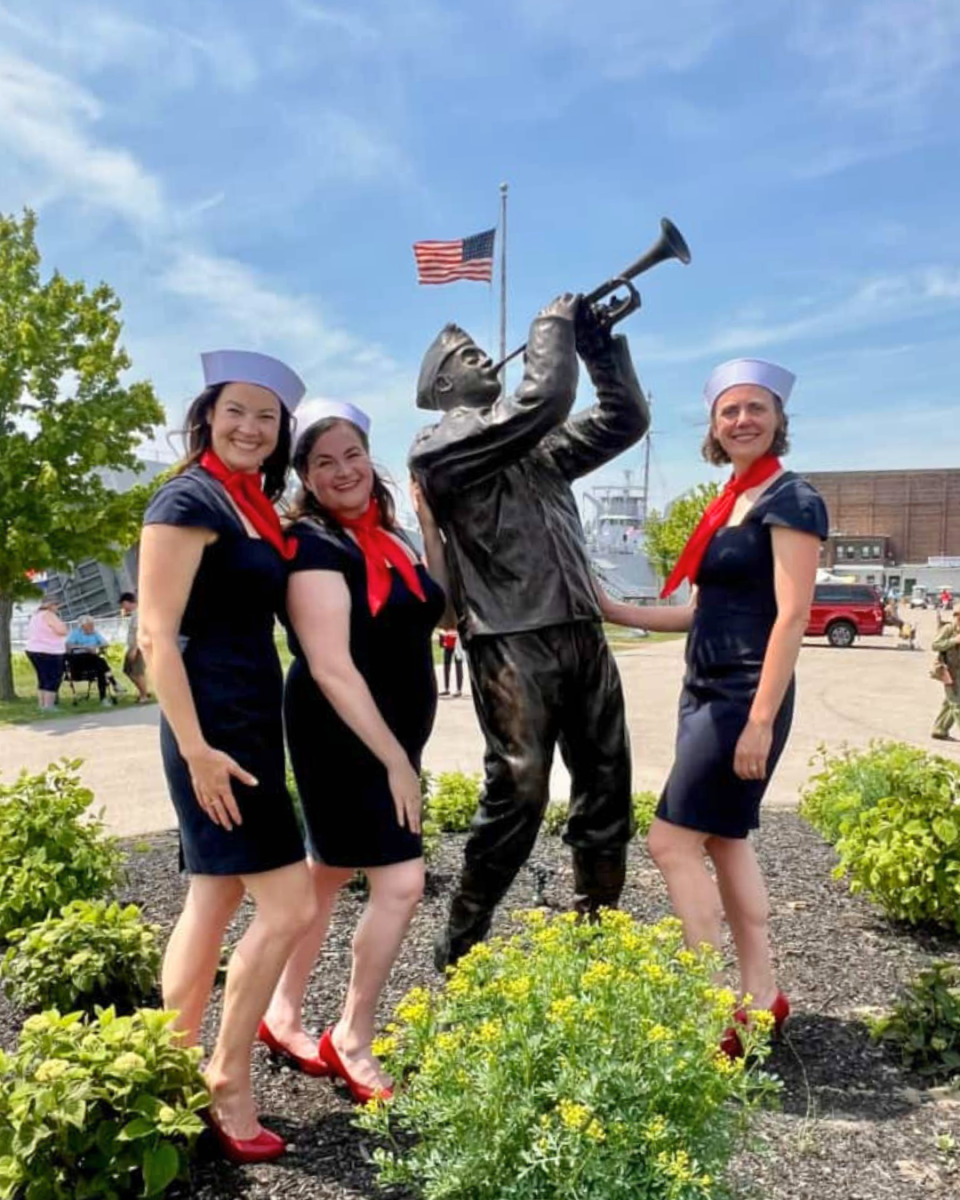 Three singers in naval themed outfits pose with a statue of a soldier playing a bugle under the American flag.