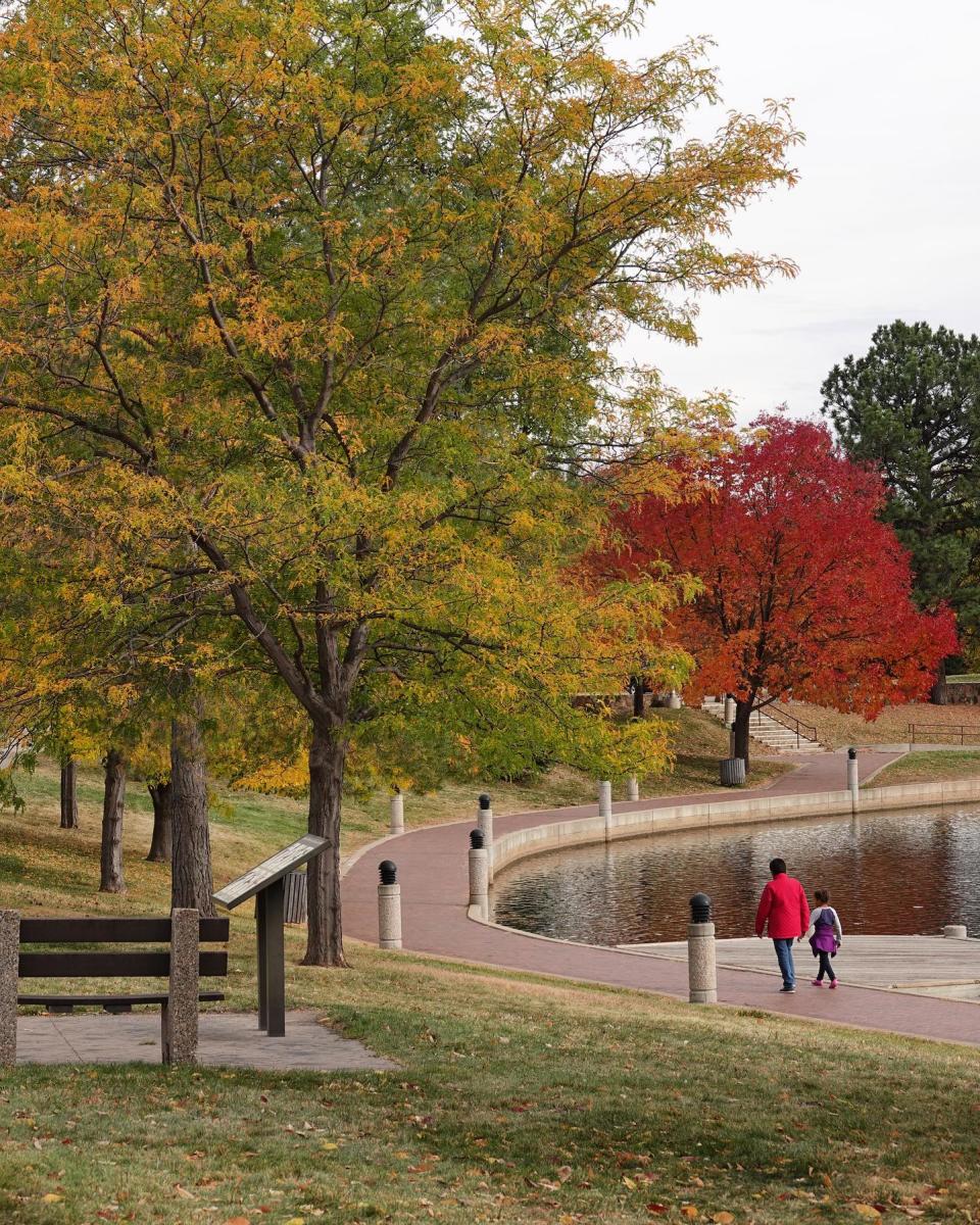 Two people walk along a curved path beside Canyon Lake water. Surrounding trees display vibrant autumn colors of green, yellow and red.