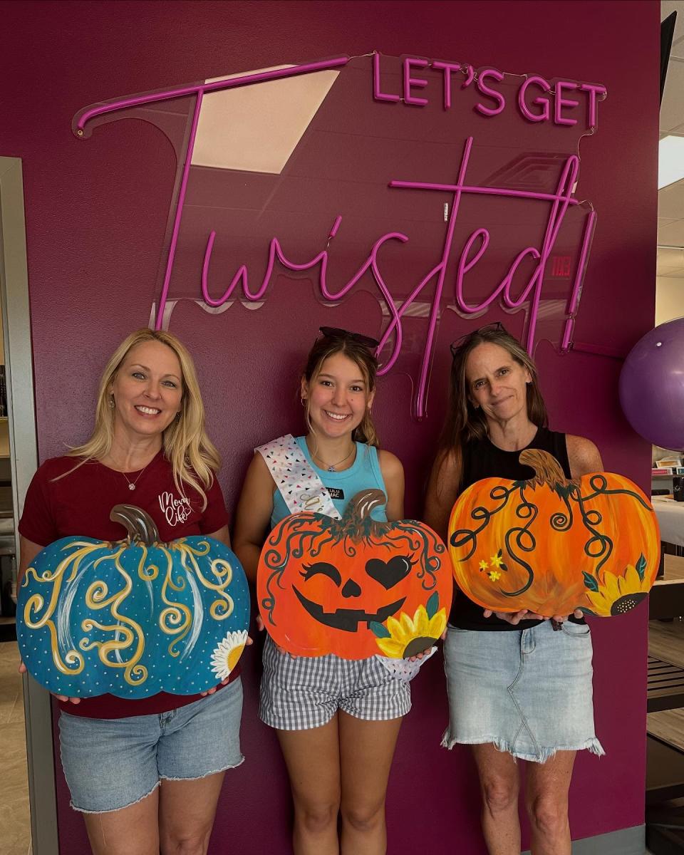 Three women hold their completed crafts--wall hangings of pumpkins painted colorfully for autumn decorating.