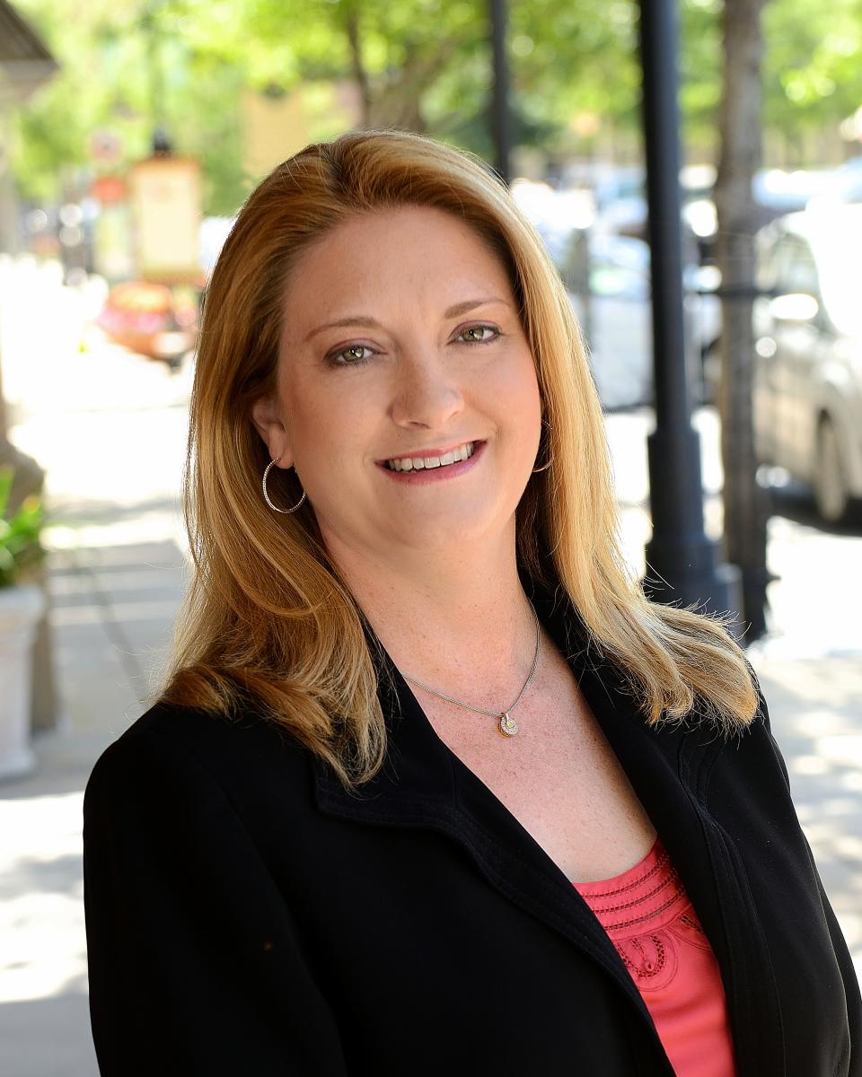A white, blonde woman smiles at the camera. She is standing outside along Market Street, wearing a coral blouse, black blazer, a small silver pendant, and small gold hoop earrings.