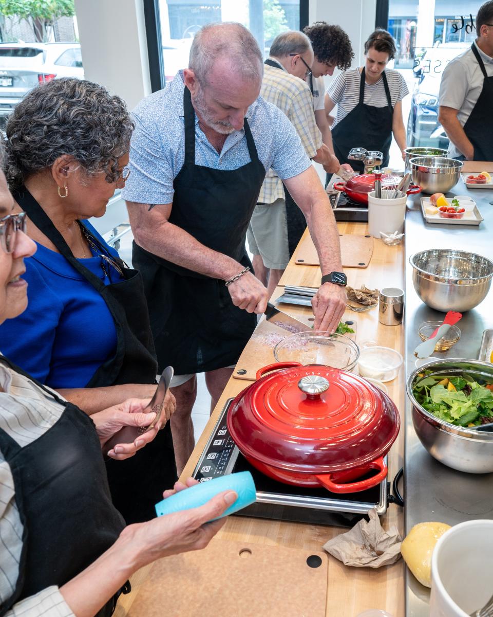 The camera looks down the length of a kitchen counter, where seven men and women of various ages and backgrounds prepare a meal as instructed by a chef (off-screen).