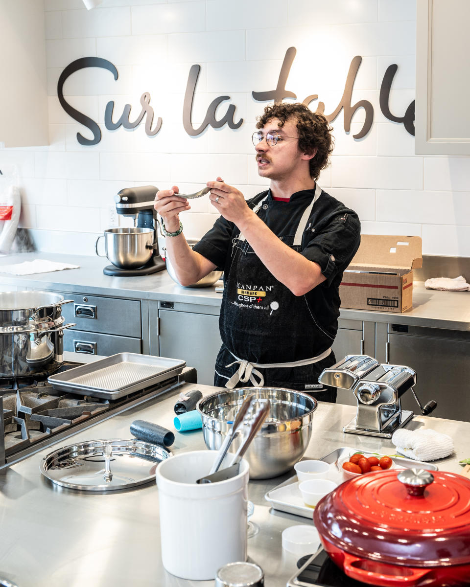 A young chef with curly brown hair, round glasses, and a black denim apron holds up a metal garlic press as he instructs a cooking class (students off screen). Various pots, pans, hot plates, and other cooking utensils are spread out on the counter before him. Behind him is "Sur la table" written in large, black, cursive.