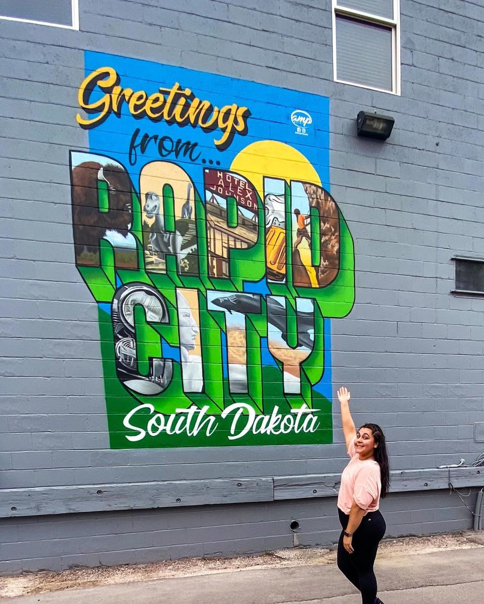 A woman in a pink shirt poses cheerfully in front of a colorful mural reading "Greetings from Rapid City, South Dakota" on a gray brick wall.