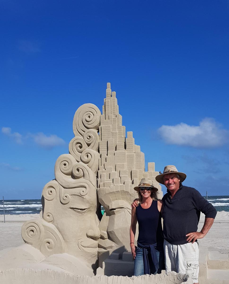 A couple wearing sun hats poses together in front of a sand sculpture showing two faces facing each other, one with a pointy top and one with a swirly top.