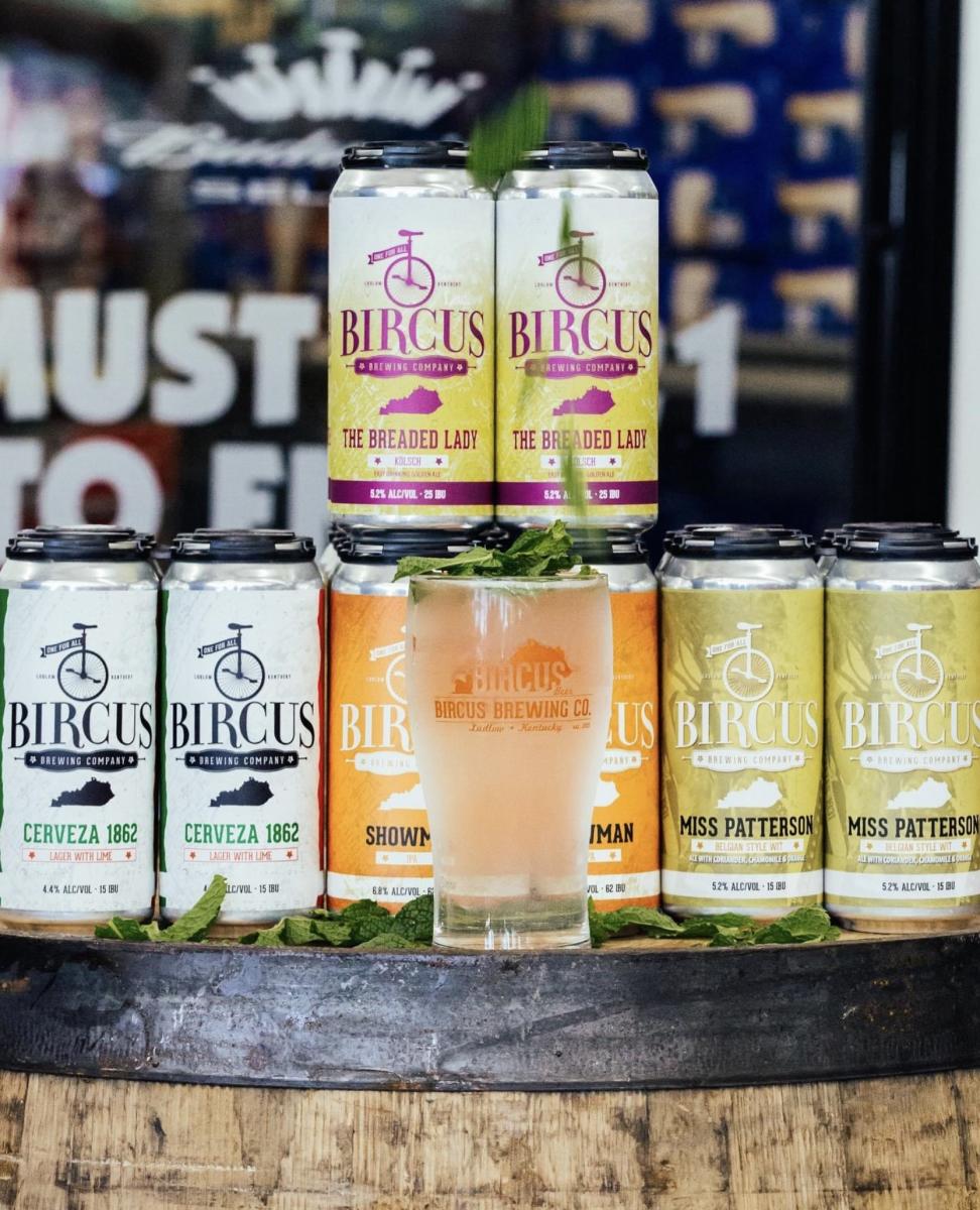 Image is of a frosted "Bricus Brewing Co." glass with cans of Bircus Brewing Co. beer behind it.