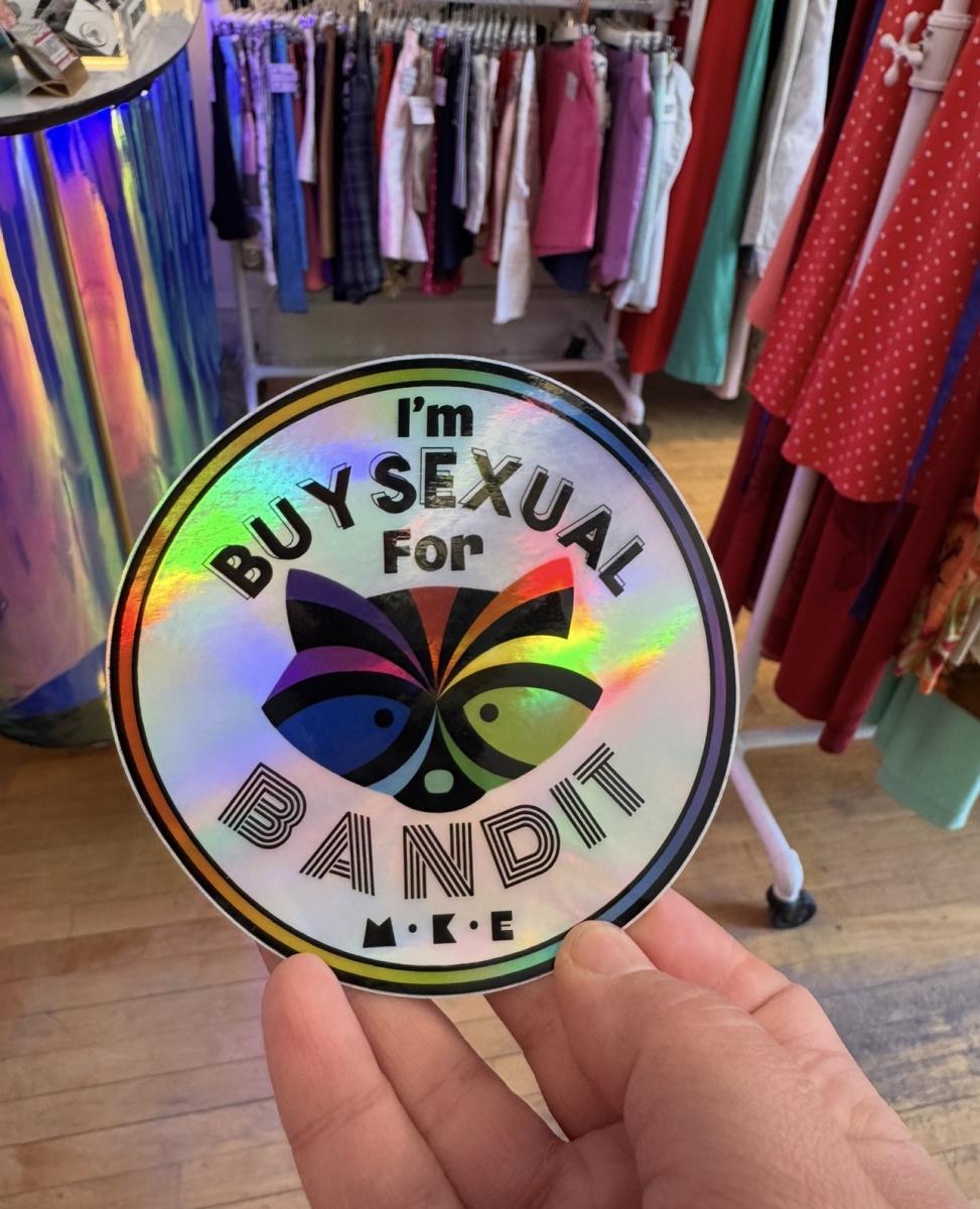 A hand holds a holographic Bandit MKE sticker reading, “I’m buysexual for Bandit,” inside the vintage shop.