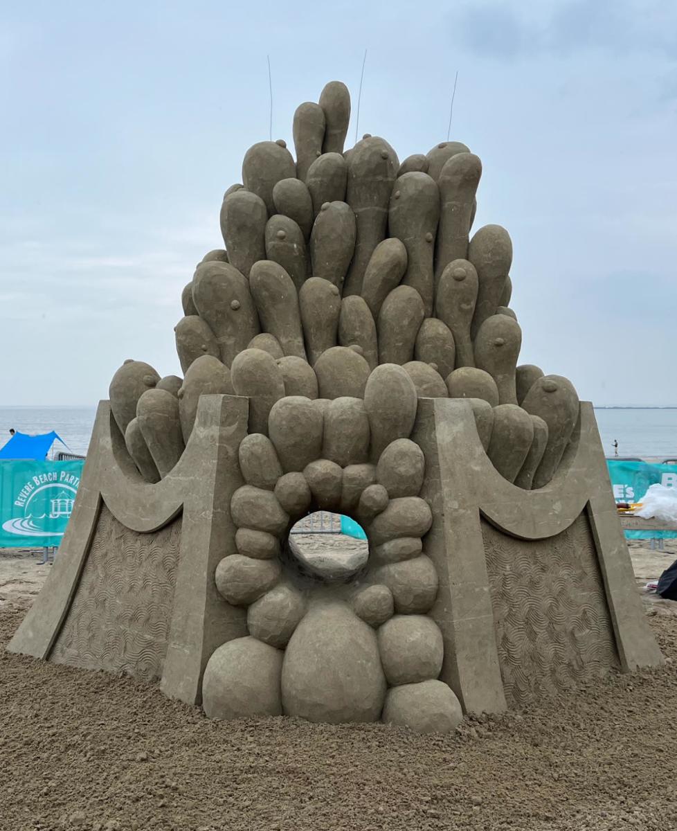 Abstract sand sculpture that has two mirrored, smooth arches on either side, with a group of bulbous shapes on the outside. There is a hole in the middle.
