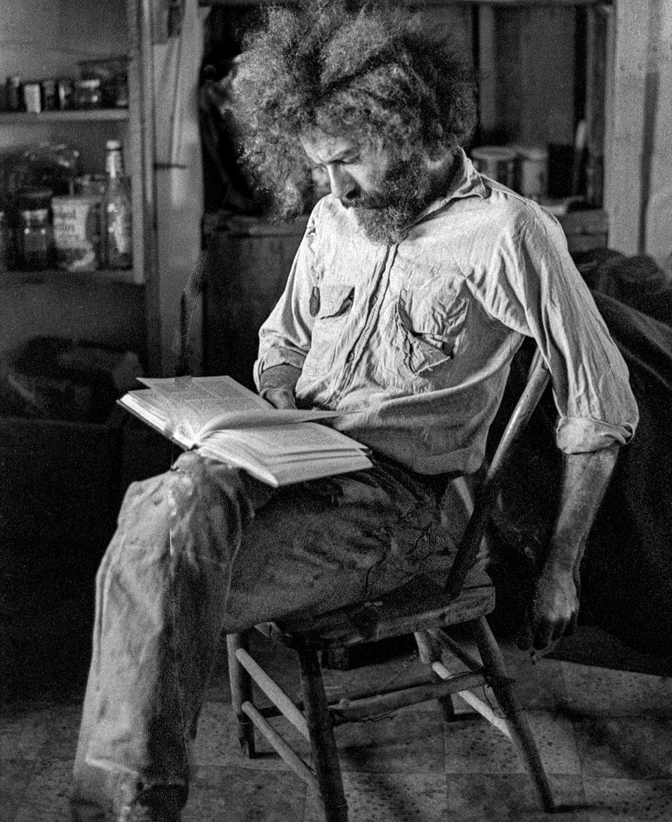 Black and white photograph of a bearded man seated indoors on a wooden chair while reading.