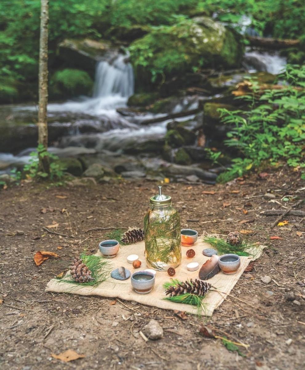 A serene outdoor tea setup beside a small forest waterfall. A burlap cloth is laid on the forest floor, topped with a glass jar filled with pine needles and water, surrounded by ceramic cups, pinecones, pebbles, and sprigs of evergreen. The background shows lush green foliage and a softly flowing creek cascading over rocks.
