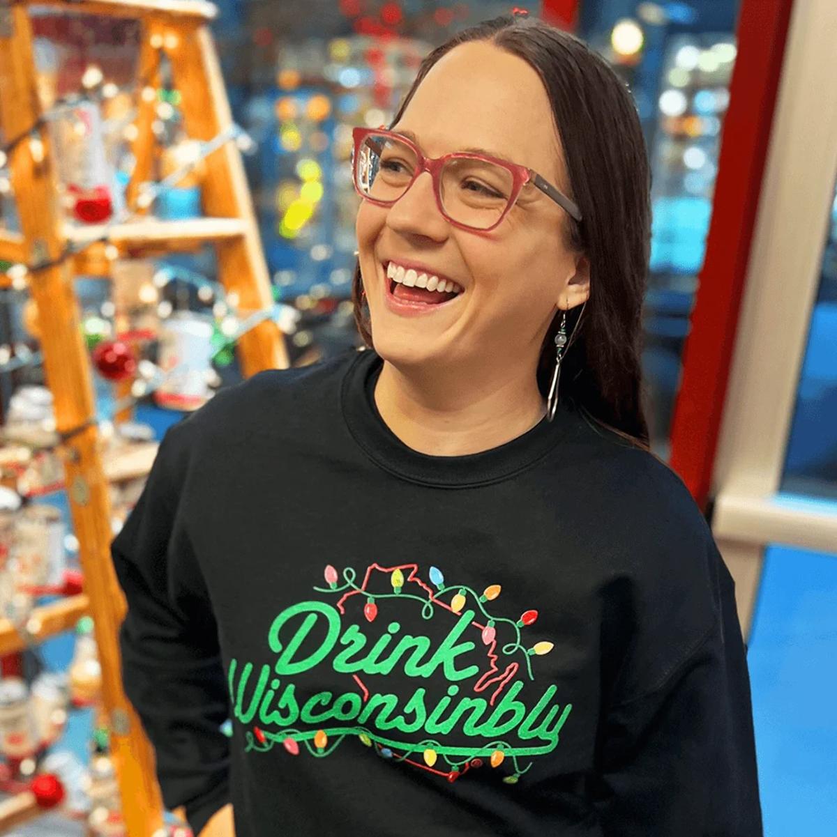 A smiling person wearing glasses and a black T-shirt that reads “Drink Wisconsibly” in colorful lettering, standing indoors with softly lit shelves and decorative items in the background. The person appears cheerful and mid-laugh in a lively, welcoming setting.