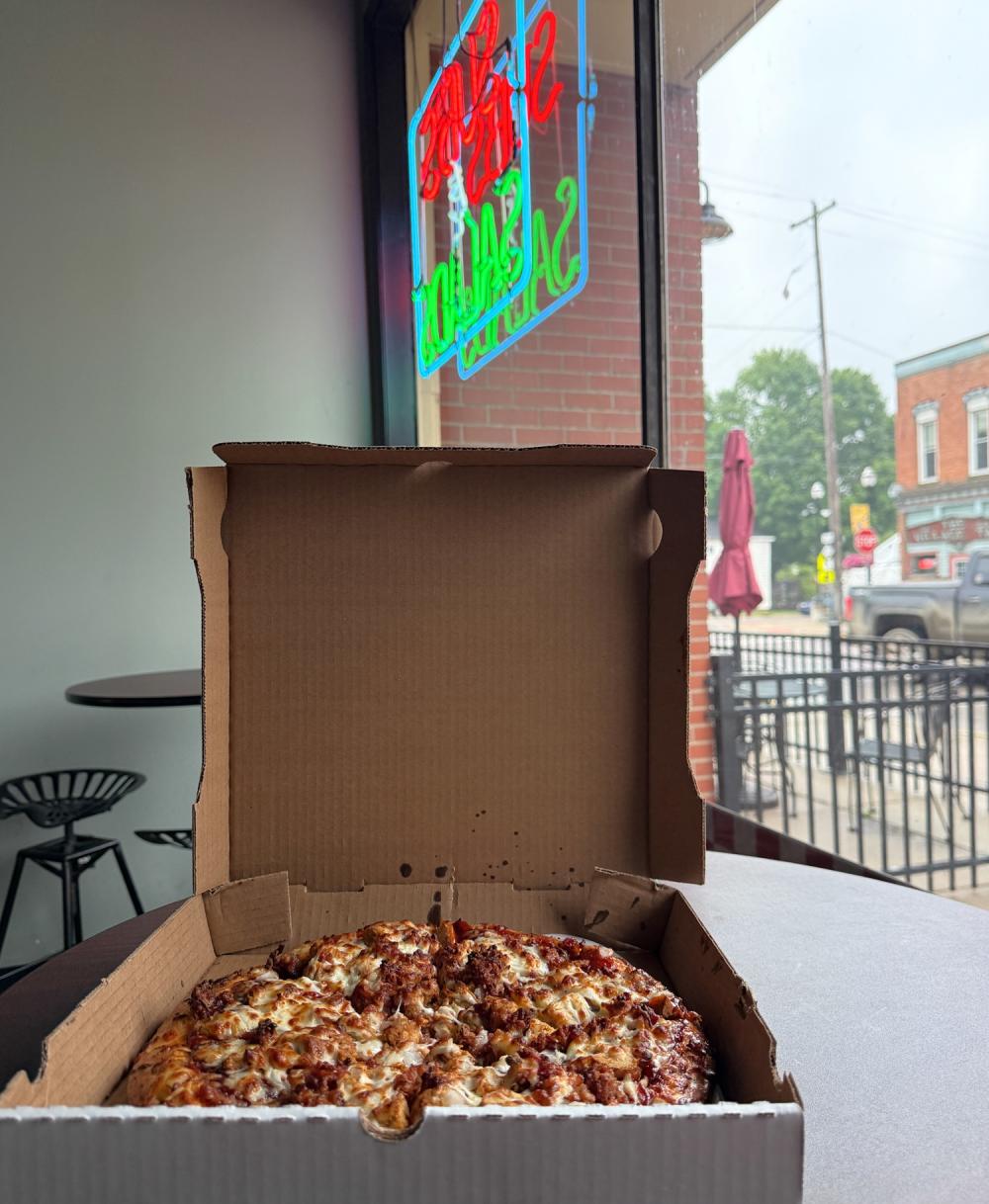 Pizza box opened with full pizza inside, sitting on a table with window facing road. Light up sign in the window facing road. Location: Ollie's pizza and grinders
