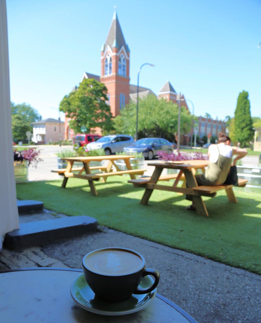 Patio view from Cafe Liv of historic church building across the street, with picnic tables on green grassy area as a person studies. Coffee sits on a table in front of the camera.