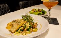 A plated trout dish with a creamy sauce and microgreens on top is next to a tall cocktail glass containing a pink drink and a smaller plate with a salad.