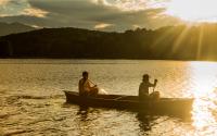 A pair of paddlers in a canoe are headed towards the right and cast in a yellow light from the setting sun, which has beams visible in the right corner of the photo.