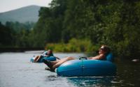 A woman relaxes in a tube with her legs hanging over the left side and her head resting on the other side. Behind her is another person in a tube with their hand in the river and a mountain in the background.