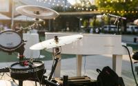 A close-up view of a white piano and black electric drum set. The venue is outdoors, and the sun is setting. There are golden light strands in the background.