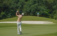 A man in a tan shirt, white pants, and a white hat swings a golf club overhead, sending the ball over a sand trap toward the hole.
