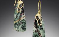 A pair of green stone earrings with small black and brown spots. The setting is gold, making two small loops around the top of the stones before forming the earring hook.