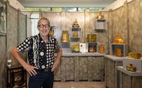 A man, 60s, smiles at the camera. He's wearing round, thick-rimmed glasses and a black shirt with large white dots and scribbles. Behind him is a booth displaying his ceramic artwork.
