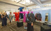 An art booth at the Texas Fine Craft Show features a prominent display of three East Asian-inspired clothing designs, one bright red, one blue, one hot pink. SuzKaz, a woman with short-cropped blonde hair, thick-rimmed glasses, and a thick animal-skin shawl, stands with her work as guests admire her work.