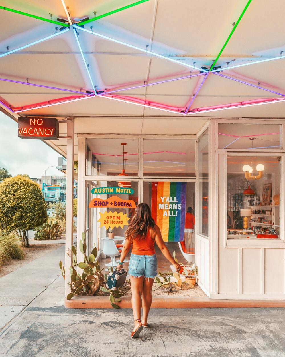 Girl walking away from the camera in front of the window at Austin Motel with a Pride flag hanging in the window with the words "Y'all Mean All".