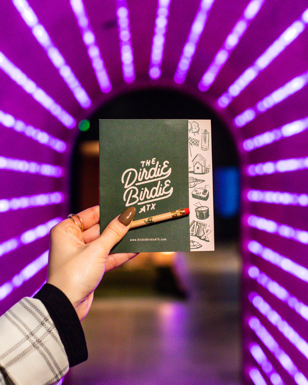 Woman's hand holding a Dirdie Birdie putt-putt score card and pencil in a purple, neon-lit tunnel.