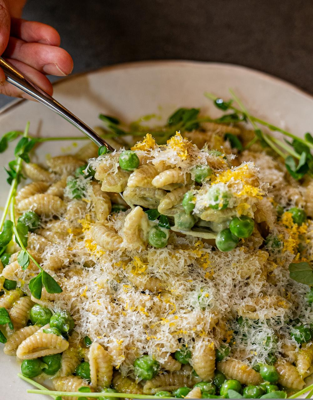 Hand scooping a spoonful of pasta covered in finely shredded cheese, peas and microgreens.