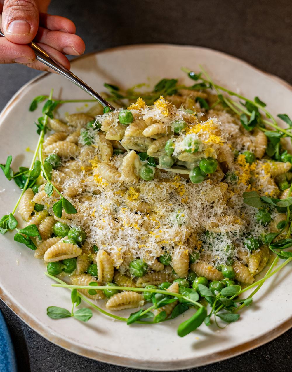 Hand scooping a spoonful of pasta covered in finely shredded cheese, peas and microgreens.