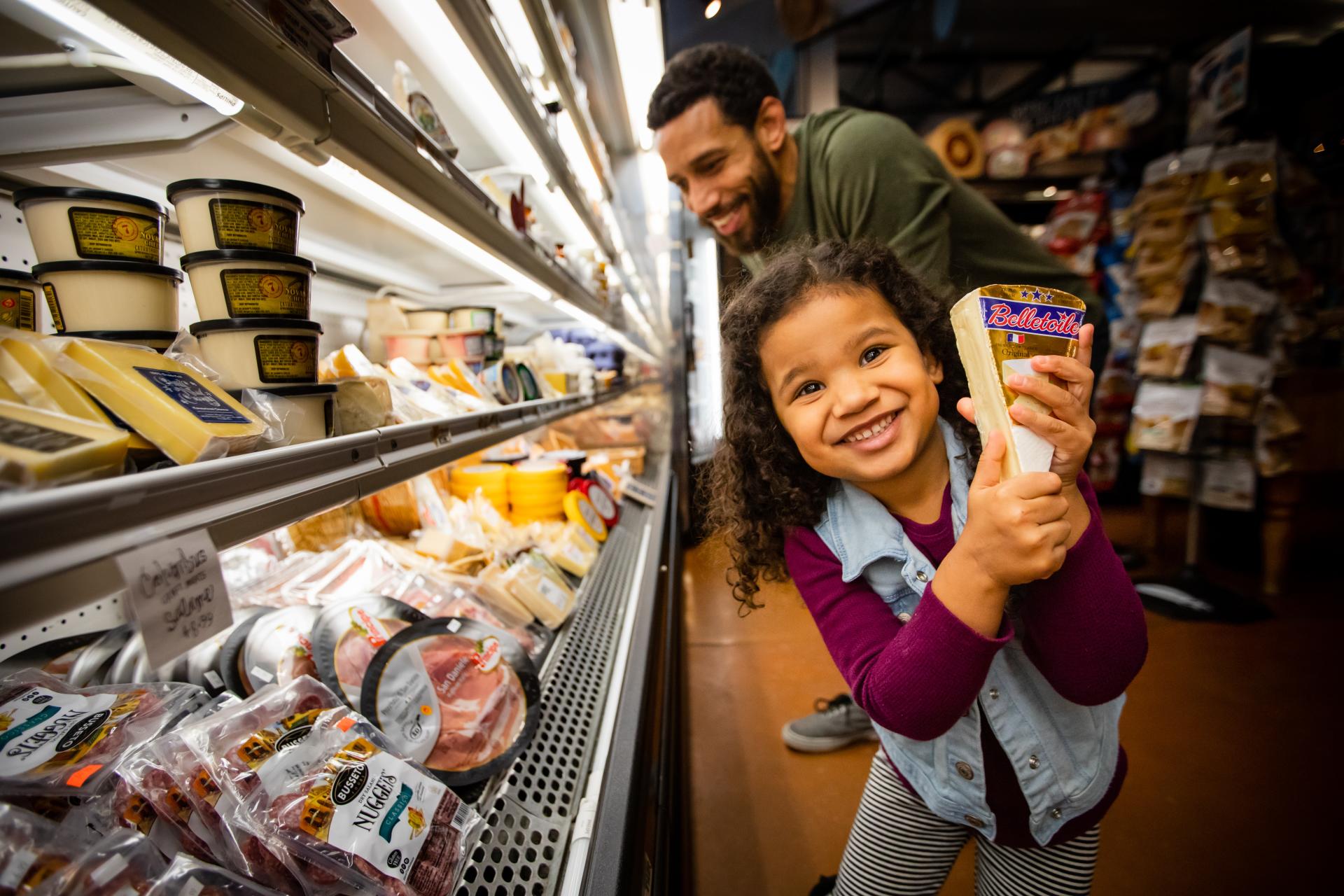 a little girl showing off a wedge of cheese in front of a large deli cooler