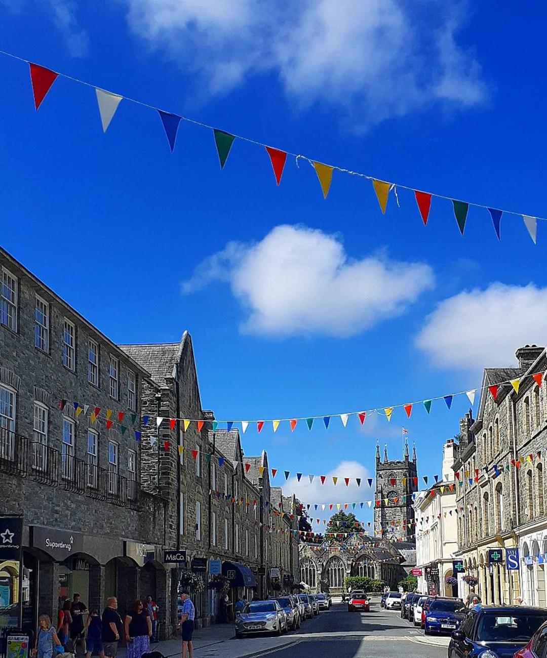 A vibrant street scene with colorful pennant flags strung between gray stone buildings under a bright blue sky. People and parked cars line the street.