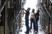 This image depicts workers in a beverage processing facility.  Three workers and inspecting tanks and water lines inside an industrial building.