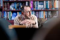 White man in glasses in a meeting with a bookcase behind him