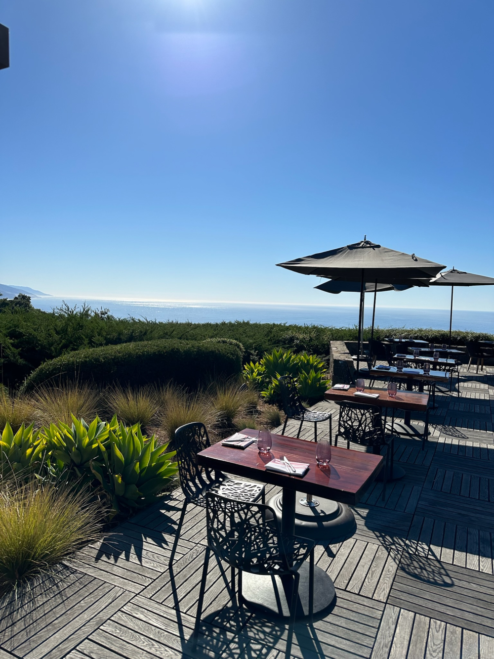 This is an image of outdoor patio tables at Alila Ventanaʻs restaurant Sur House in Big Sur. The patio tables are shaded by umbrellas. The bright blue ocean can be seen in the background