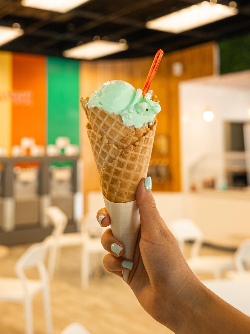 A young woman with light blue nails holds up a waffle cone filled with light green (pistachio?) ice cream. The background (out of focus) features a wall painted with thick vertical stripes in yellow, orange, and bright green. Below the stripes is a row of froyo dispensers, all labeled with their flavors. There are several small round table with white plastic chairs in the middle of the room.