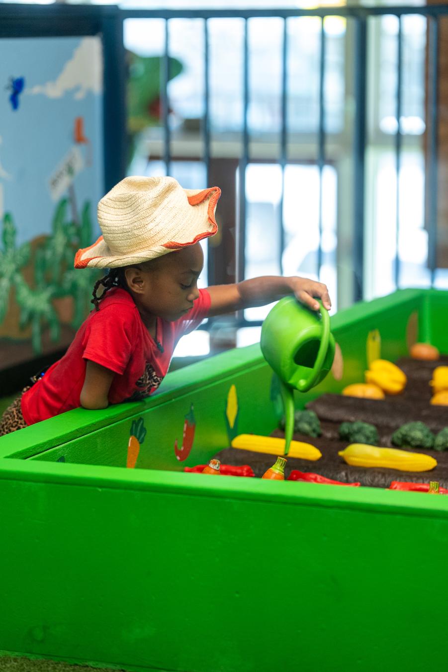 Little girl wearing a red shirt and a straw hat watering a play garden at Imagination Place.