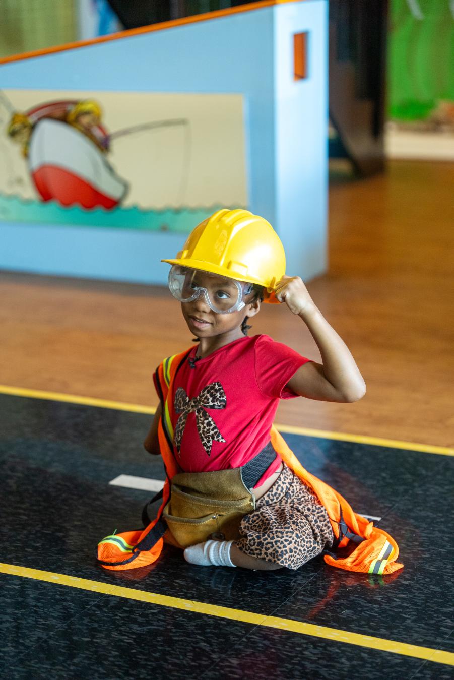 A young girl wearing a construction hat and vest sitting in the construction play zone at the Imagination Place.