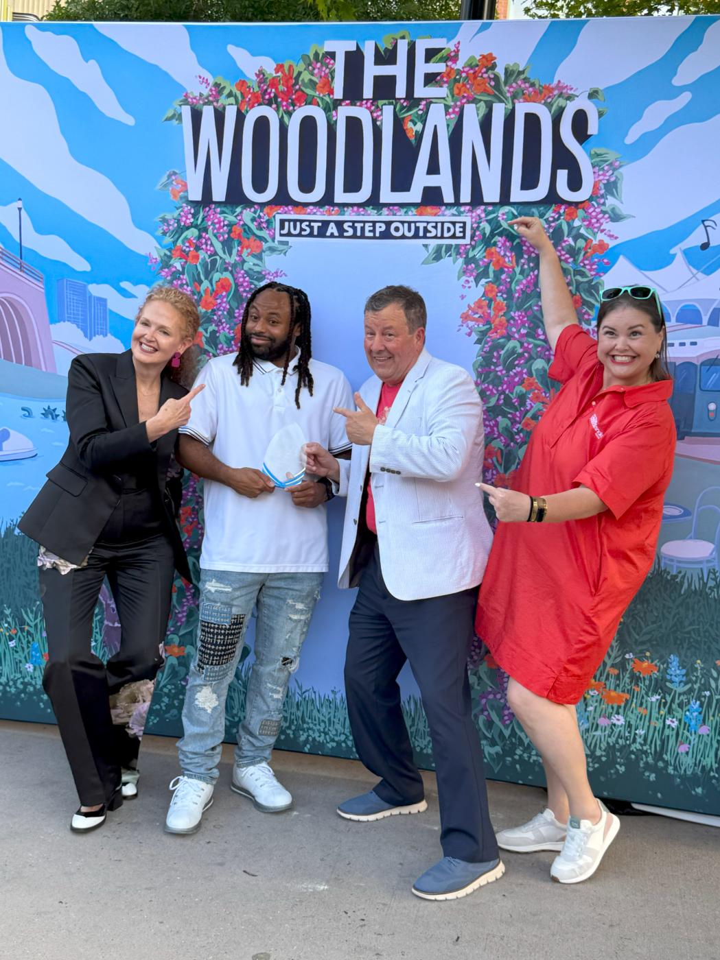 Four people pose for the camera: Kim Lowe (curly red hair, classy black pantsuit), Sterling Scott (dreads, white polo, distressed jeans), Nick Wolda (red T-shirt, white blazer, navy slacks, denim sneakers), and Elizabeth Eddins (bright red sundress with teal sunglasses on her head). Sterling Scott is holding a clear glass award, which he received for his excellence in tourism service as a valet at The Woodlands Resort. The group is standing in front of a backdrop depicting a colorful, stylized mural of The Woodlands.