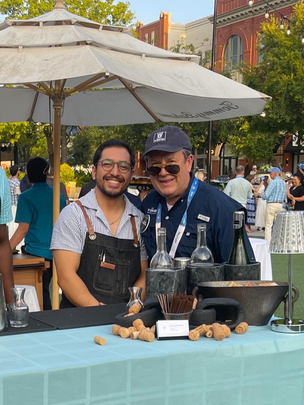 Jose Perez (left, wearing a striped polo and dark denim apron) and Nick Wolda (wearing a navy sport shirt and ballcap with the "Visit The Woodlands" old logo), smile at the camera. They're standing behind a wine station at the TSAE opening reception, catered by Sixty Vines, and under a quality Tommy Bahama patio umbrella.