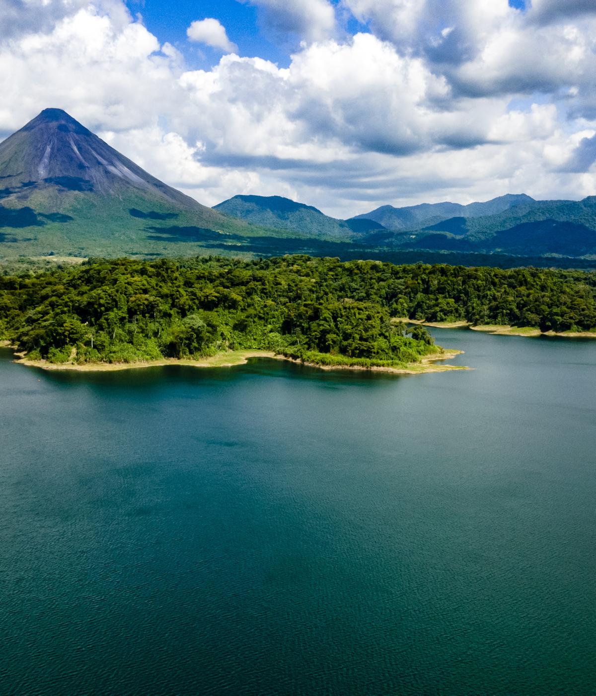 An aerial view of Lake Arenal, Costa Rica, with the Arenal Volcano in the background. The volcano  is a cone-shaped peak with clouds around its summit. The lake is surrounded by lush green  rainforest and a dam is visible on the left side of the image.