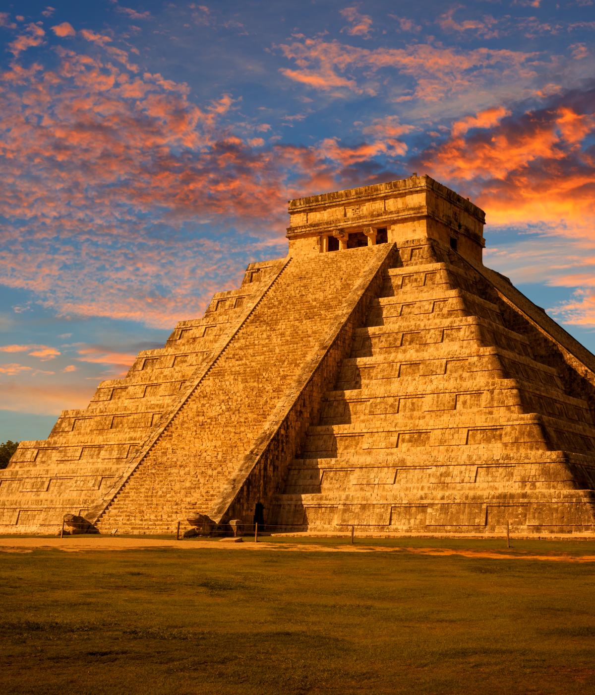 The iconic Temple of Kukulkan, at Chichen Itza in Mexico. The pyramid is bathed in  golden light against a dramatic sunset sky.