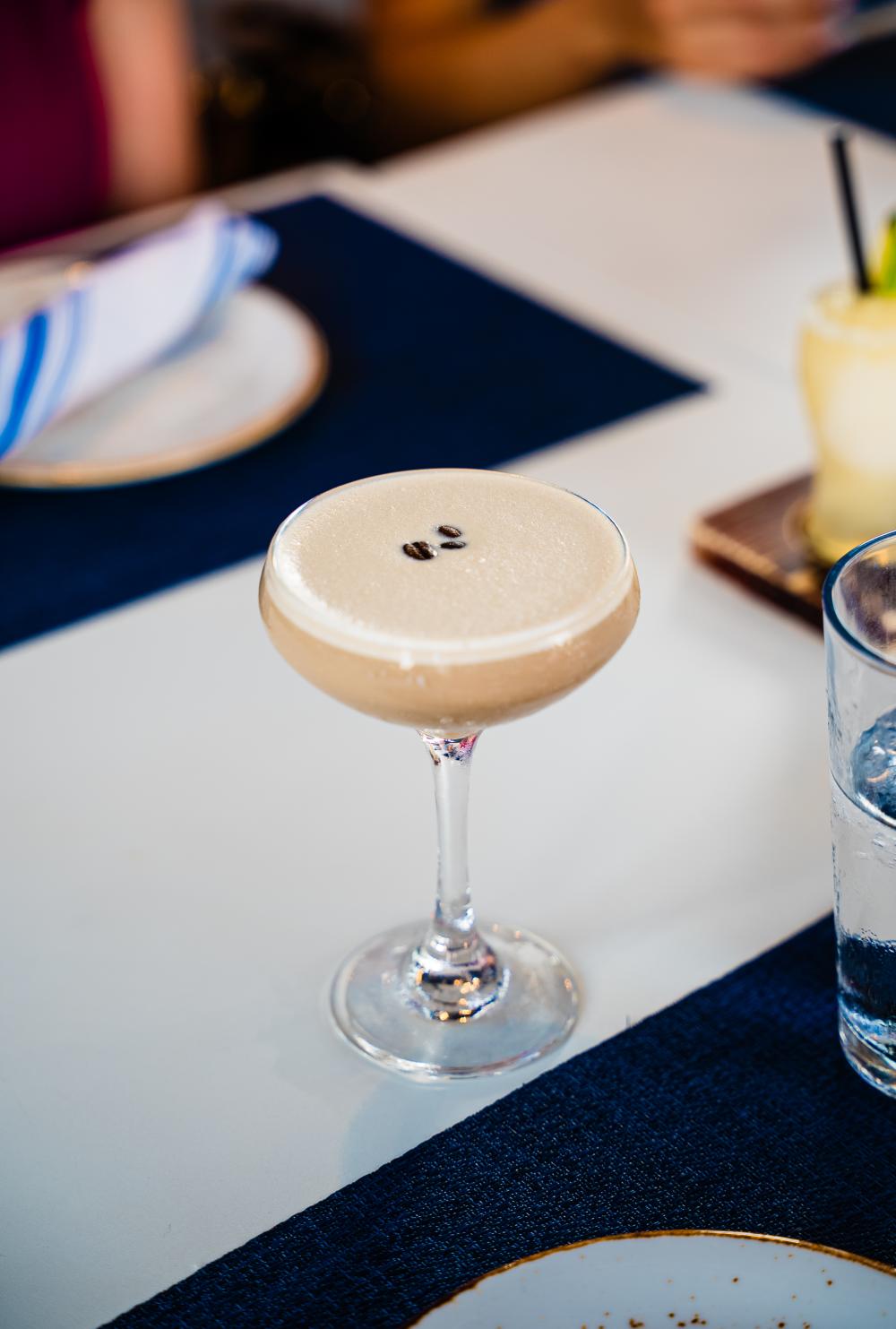 A martini glass rests on a table. The drink is a pale coffee color with three coffee beans floating on the surface. The table is set with plates, cloth-napkin-wrapped silverware, and navy placemats. Off to the right are a glass of water and the corner of a margarita flight.