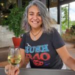 Claudia Alarcón Headshot. She is sitting at a patio table and holds a glass of white wine