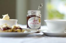 An Open pot of Tiptree Little Scarlet Jam sits, opened, beside scones, cream and a cup of tea on a pristine tablecloth.