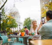 A man takes a picture of a woman with the Capitol building in the background as they enjoy sipping on coffee outside of Ancora coffee shop on King Street in downtown Madison