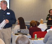 A man and woman give a PowerPoint presentation while others seated listen and take notes
