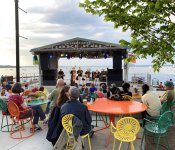 Dozens of people watching a concert at Memorial Union Terrace, sitting on the popular orange, yellow, and green terrace chairs. The people are facing the stage which is in front of Lake Mendota in the background on a sunny Summer day.
